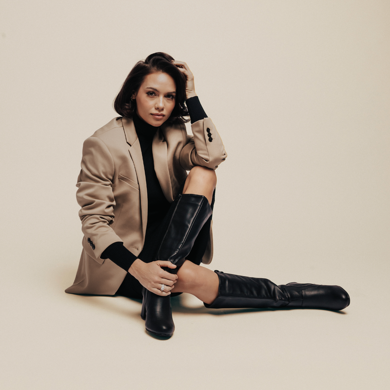 Woman sitting on the floor wearing black knee-high boots, a black turtleneck, and a beige blazer.