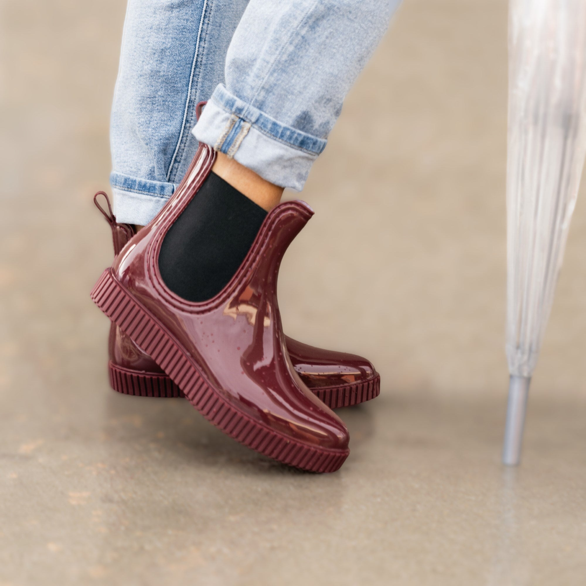 Close-up of a pair of glossy maroon slip-on rain boots with ribbed soles, styled with cuffed light-wash jeans.