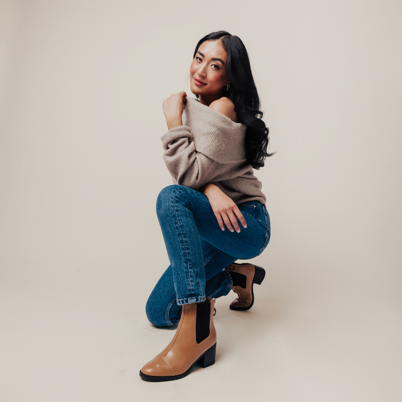 Woman wearing tan Chelsea boots, blue jeans, and an off-the-shoulder beige sweater, posing in a relaxed, kneeling position.