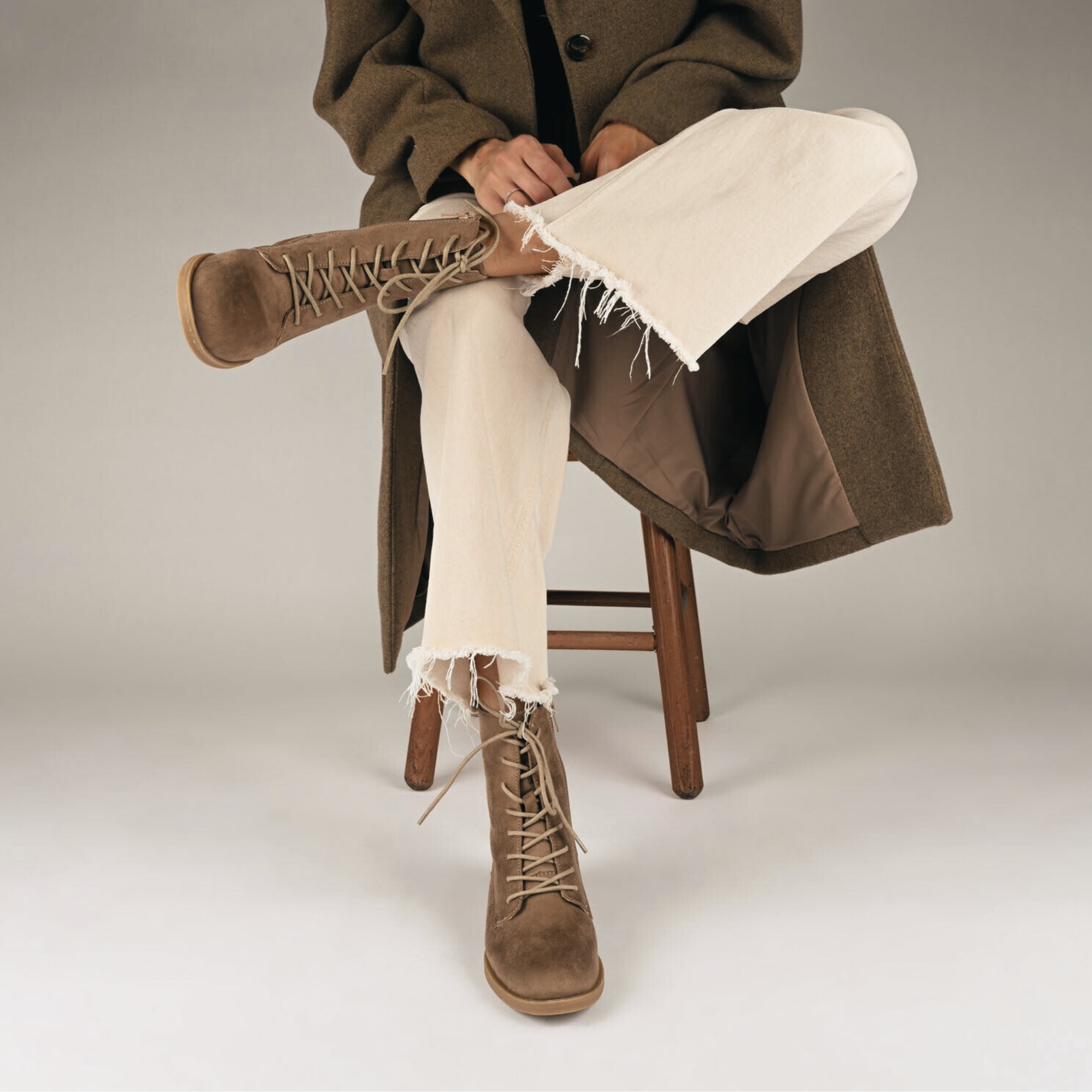 Person seated with legs crossed, wearing beige lace-up combat boots, cream-colored frayed hem jeans, and a brown coat.