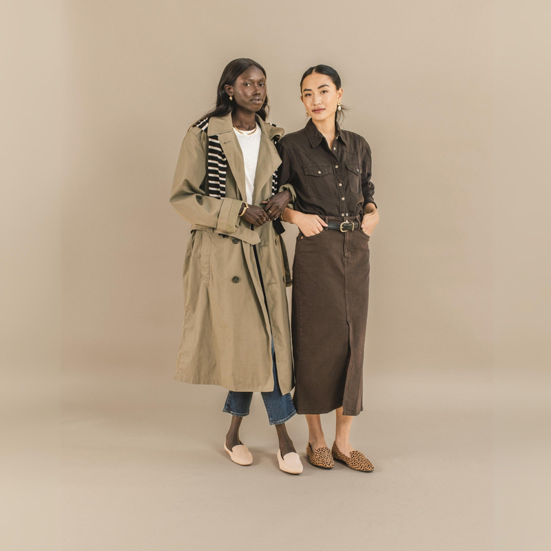 Two women standing together in stylish outfits: one wearing a trench coat, striped scarf, jeans, and light-colored loafers, and the other in a brown button-up shirt, long skirt, and leopard-print flats.