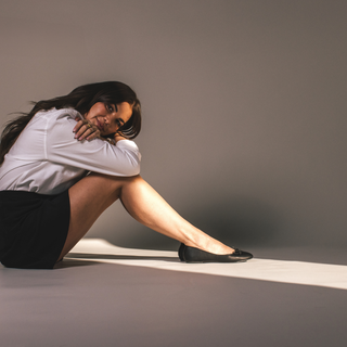 Woman sitting on the floor in a relaxed pose, wearing black ballet flats, a white shirt, and a black skirt.