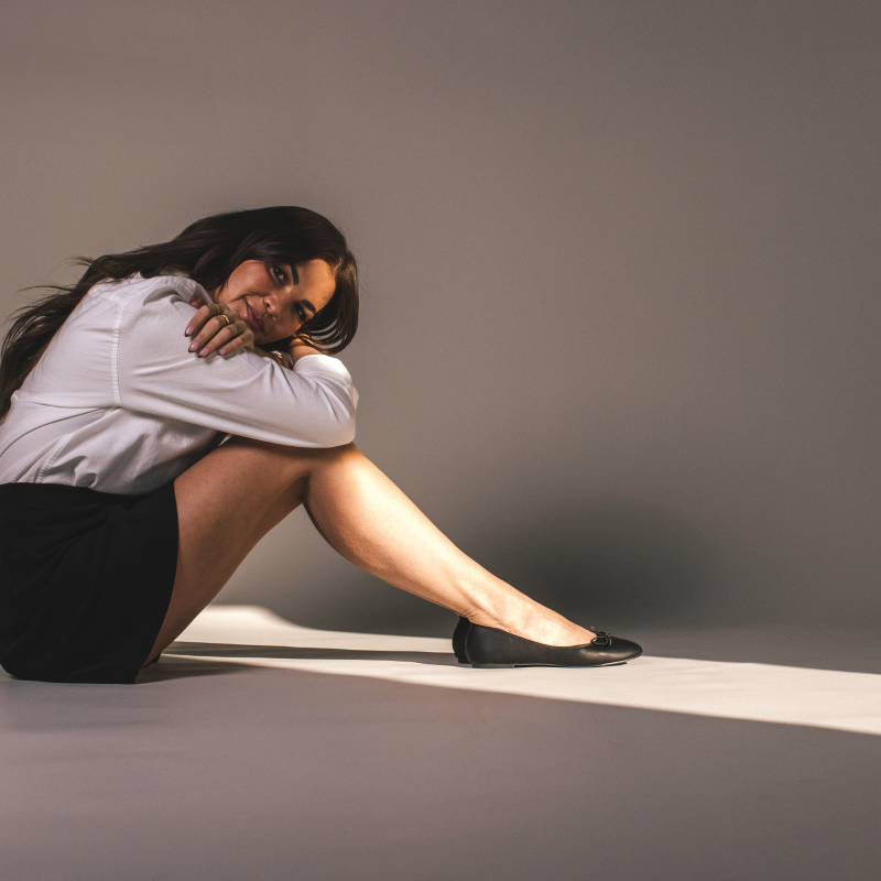 Woman sitting on the floor in a relaxed pose, wearing black ballet flats, a white shirt, and a black skirt.