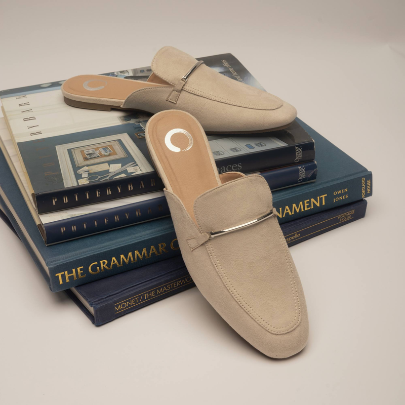 Pair of beige suede loafers with silver hardware, styled on a stack of grammar and design books.