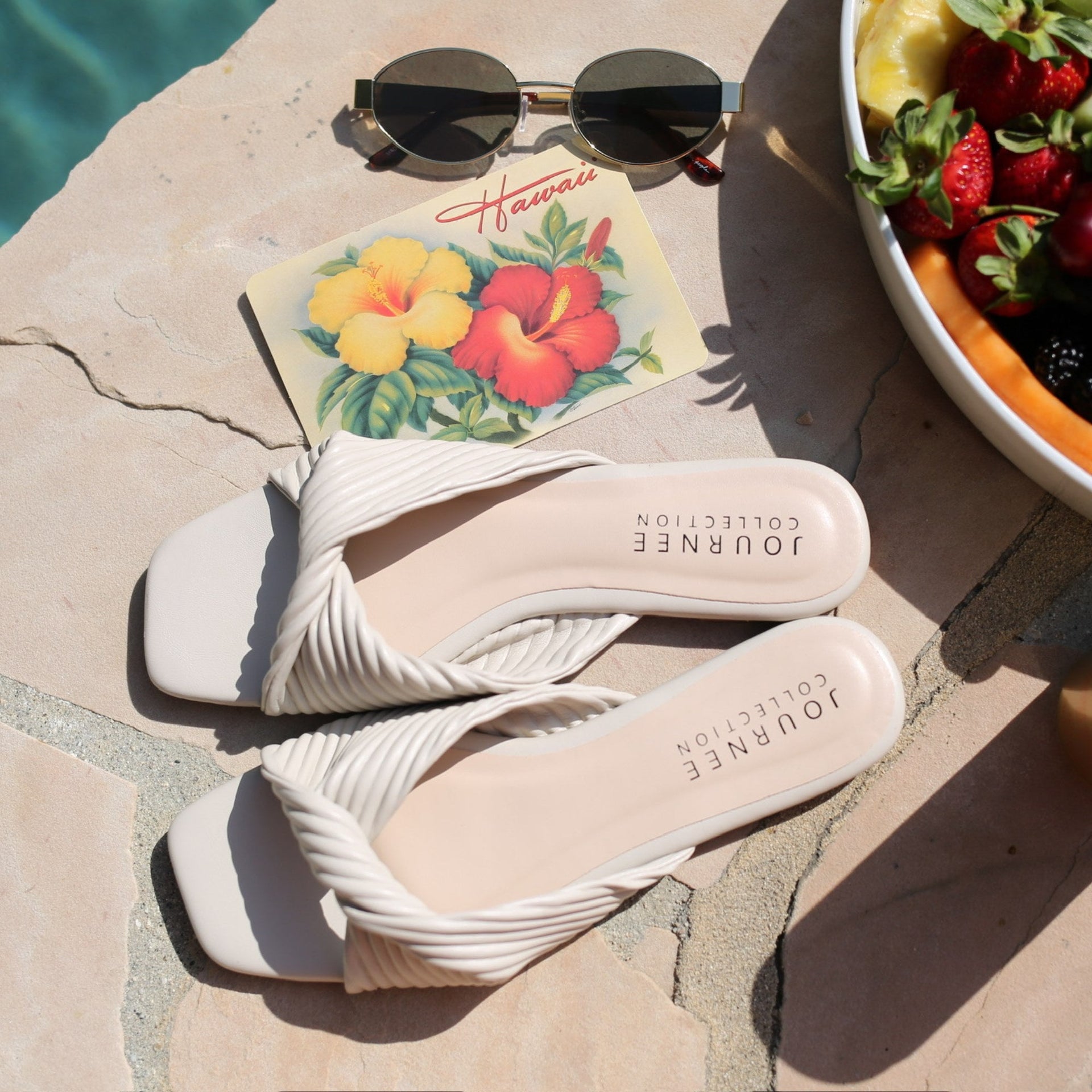 Pair of white pleated sandals from Journee placed poolside, styled with sunglasses, a postcard reading 'Hawaii' with hibiscus flowers, and a bowl of fresh fruit.