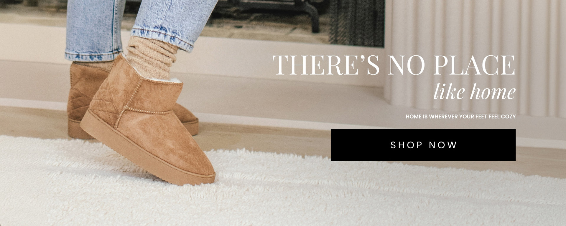 Close-up of a woman’s feet wearing cozy, tan faux suede slipper booties with a plush lining. She pairs the boots with light-wash jeans and wool socks, standing on a soft, white rug. Text overlay reads: 'There’s No Place Like Home - Home is Wherever Your Feet Feel Cozy.' Shop Now button included.