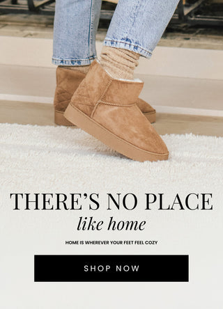 Close-up of a woman’s feet wearing cozy, tan faux suede slipper booties with a plush lining. She pairs the boots with light-wash jeans and wool socks, standing on a soft, white rug. Text overlay reads: 'There’s No Place Like Home - Home is Wherever Your Feet Feel Cozy.' Shop Now button included.