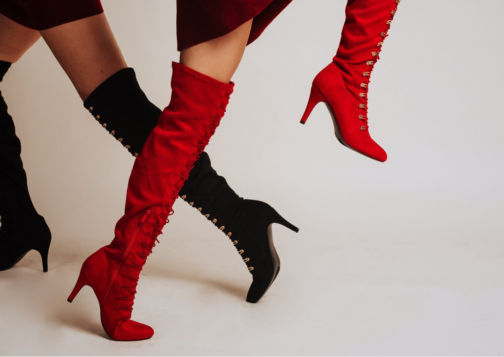 Two people wearing knee-high heeled boots with lace-up detailing, one medium calf in bold red and the other wide calf in black.