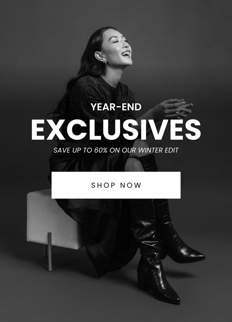 A sleek monochromatic banner featuring a smiling woman in stylish winter attire, including a glittery top, leather skirt, and tall black boots. The text reads, 'Year-End Exclusives - Save up to 60% on our Winter Edit,' with a 'Shop Now' button below.